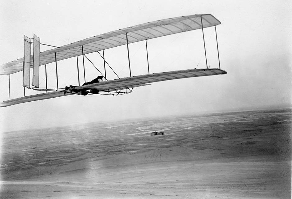Wilbur Wright flying his and Orville Wright’s 1902 glider at Kitty Hawk, North Carolina, with the brothers’ camp and shed visible in the distance, 1903