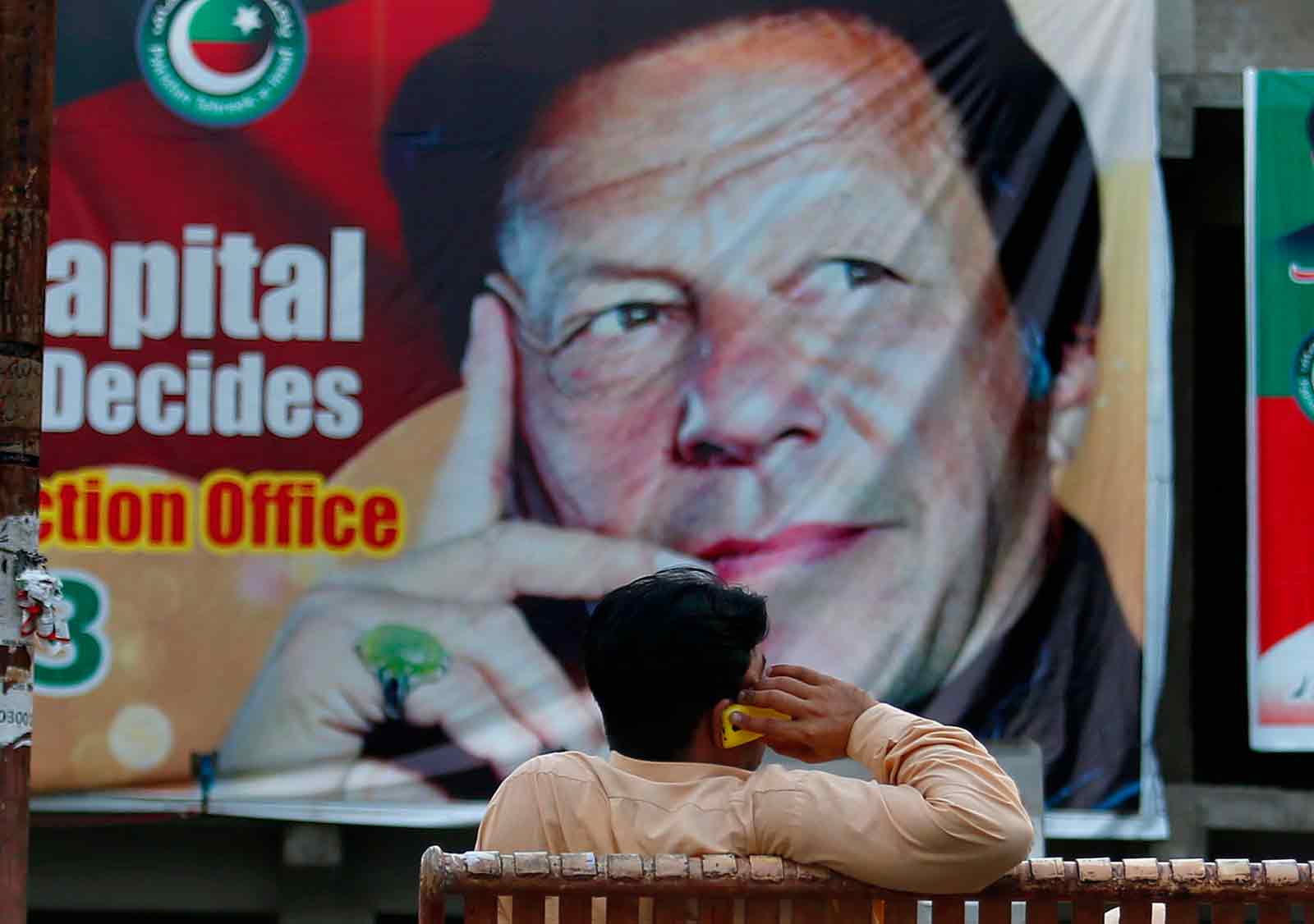 A man on his cell phone sitting in front of a poster showing Imran Khan, head of the Pakistan Tehreek-e-Insaf party, which won this past week’s elections, Islamabad, Pakistan, July 28, 2018