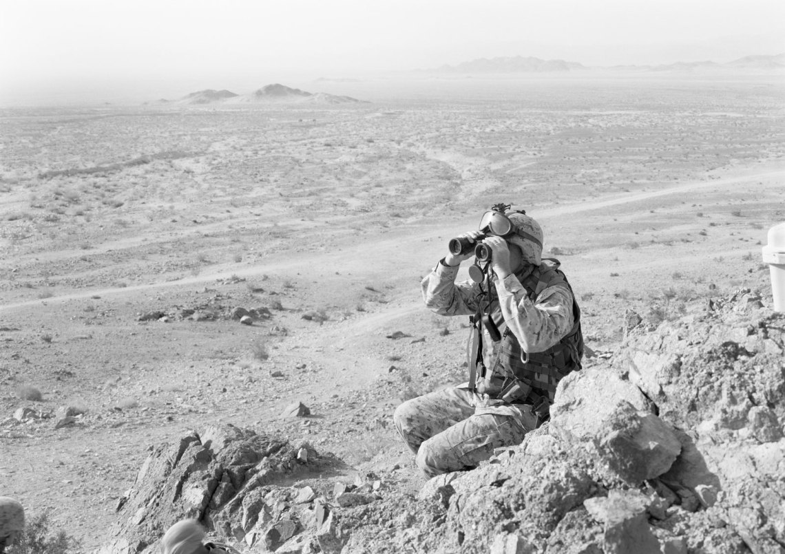 A soldier using binoculars to look out over a hilly area