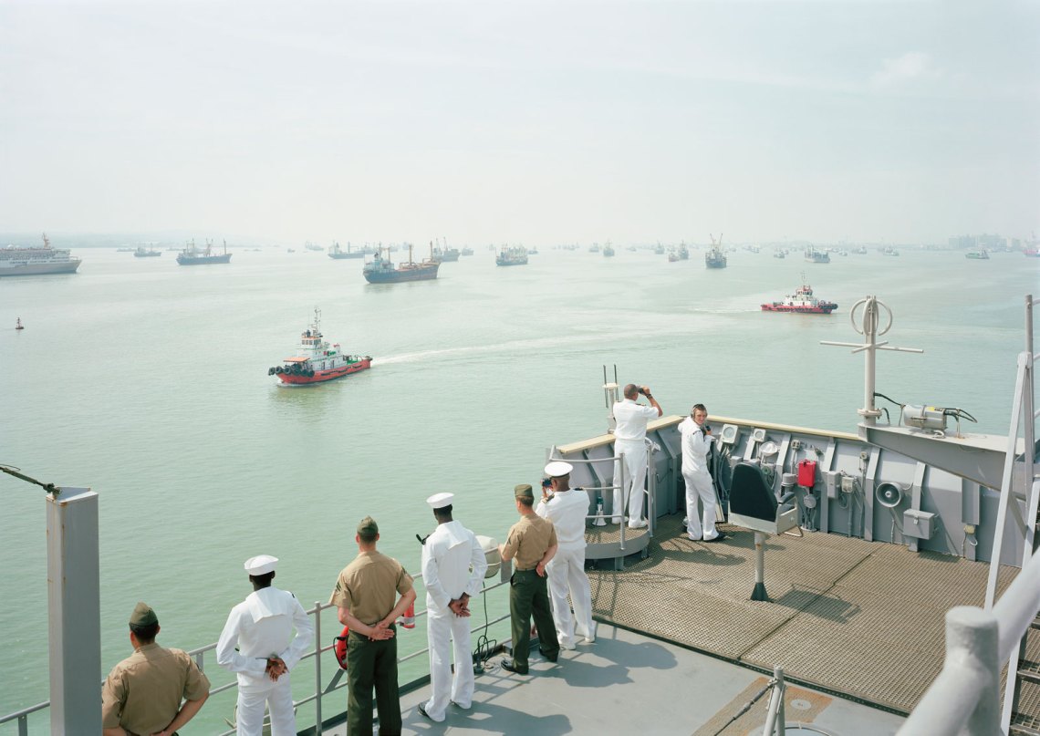 Sailors looking from the dek of a ship out onto the water, where other ships are parked
