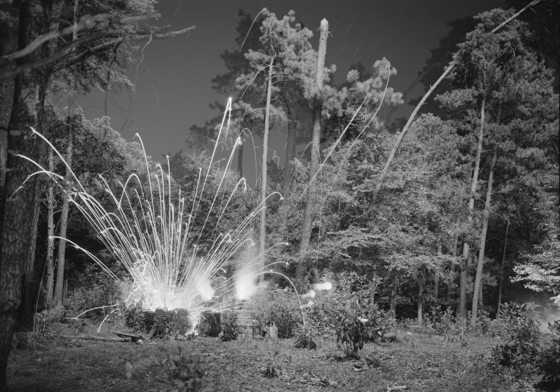 An explosion creates blasts of light in a wooded area in black and white