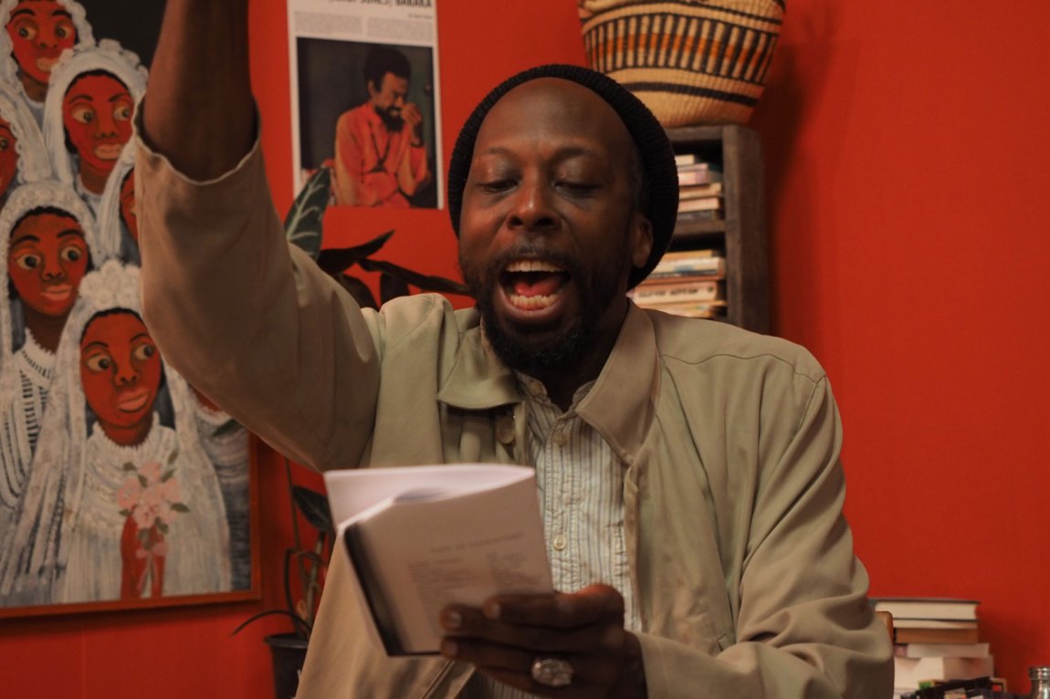 A black man reading from a text with his hand up in emphasis, before a red background
