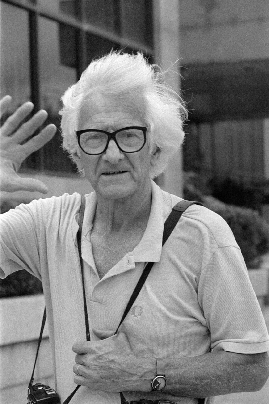 Marc Riboud, with white hair and black-rimmed glasses, waving with one hand up, the other holding the strap of the camera that hangs around his neck