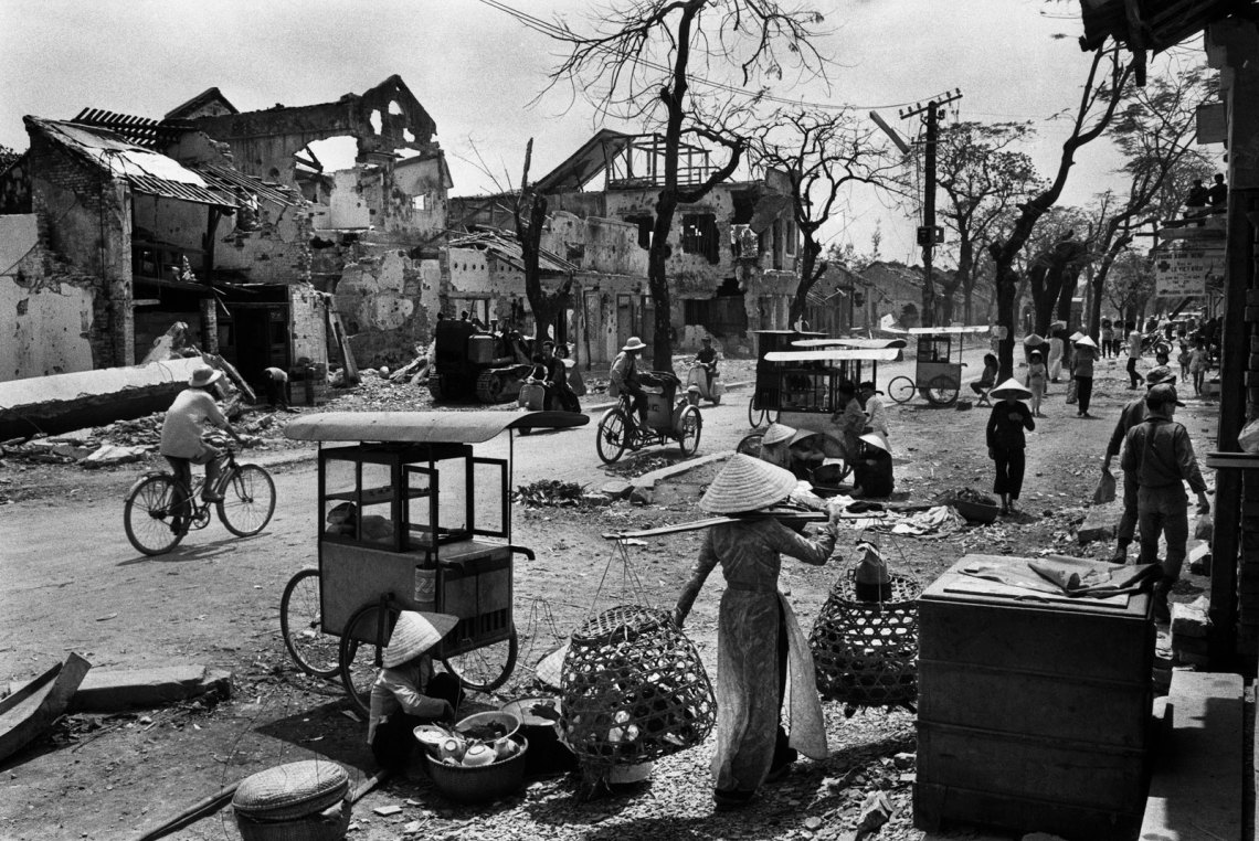 black and white image of the rubble in Hue, with people walking and bicycling, one woman in a hat carrying balanced bags over her shoulder