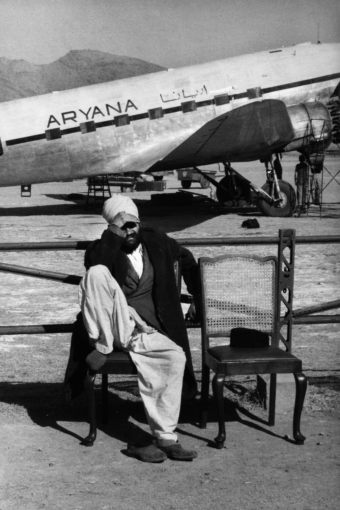 a man sitting on a chair in front of a plane, shields in face from the sun, in black and white