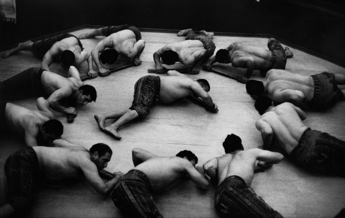 shirtless men lying on the ground form a circle, with one man in the middle, in black and white