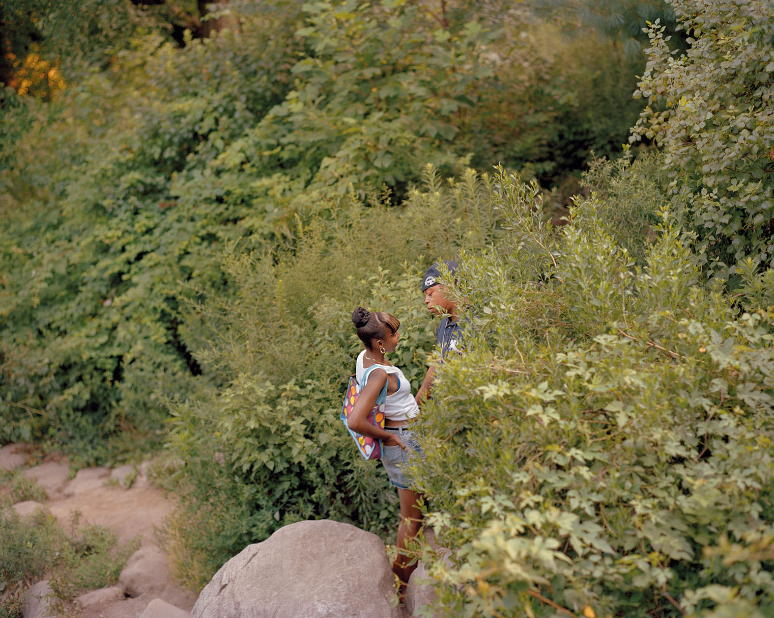 A couple talks, partially obscured by shrubs, in a green and overgrown area of the park