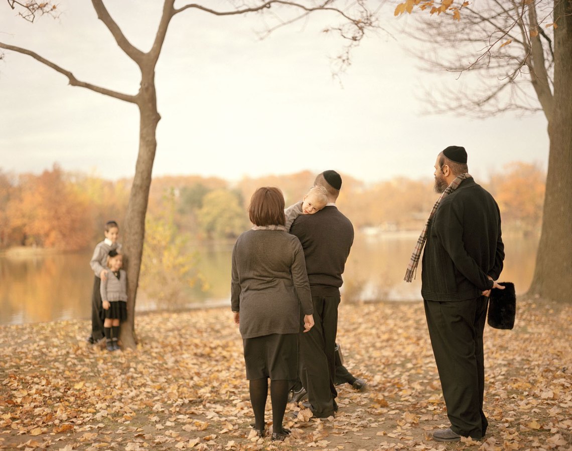 a Hasidic family stands, dressed nicely, on an autumn day, two children posed by a tree, three adults, one carrying a baby, looking on