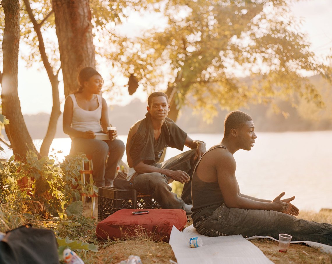 three African American figures, a woman and two men, sit near the lake, all awash in golden light and two men