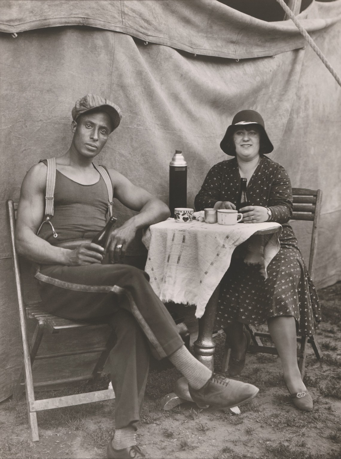 Two circus performers sit at a table in front of a tent, drinking out of a thermos
