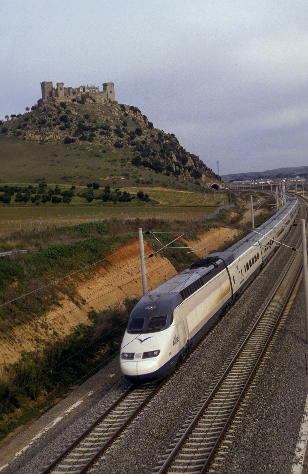 A high-speed train comes down the track around an outcropping with the ruins of a castle