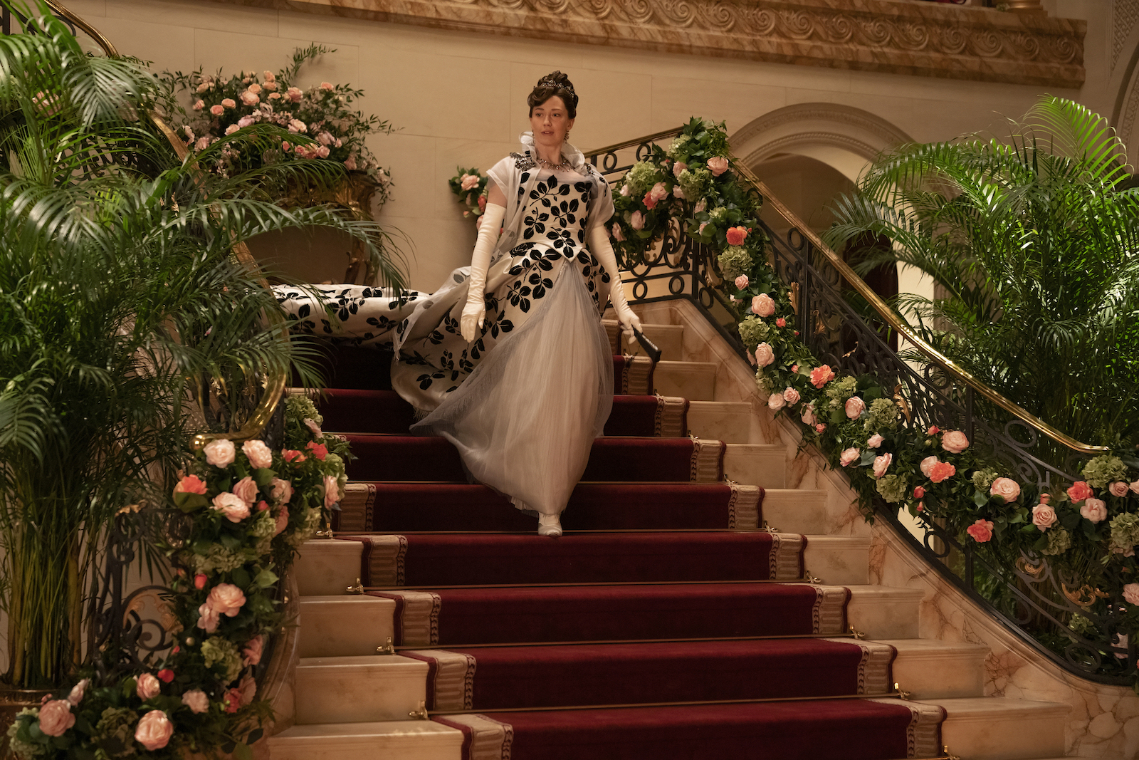 A woman in Gilded Age finery descends the stairs of her ballroom