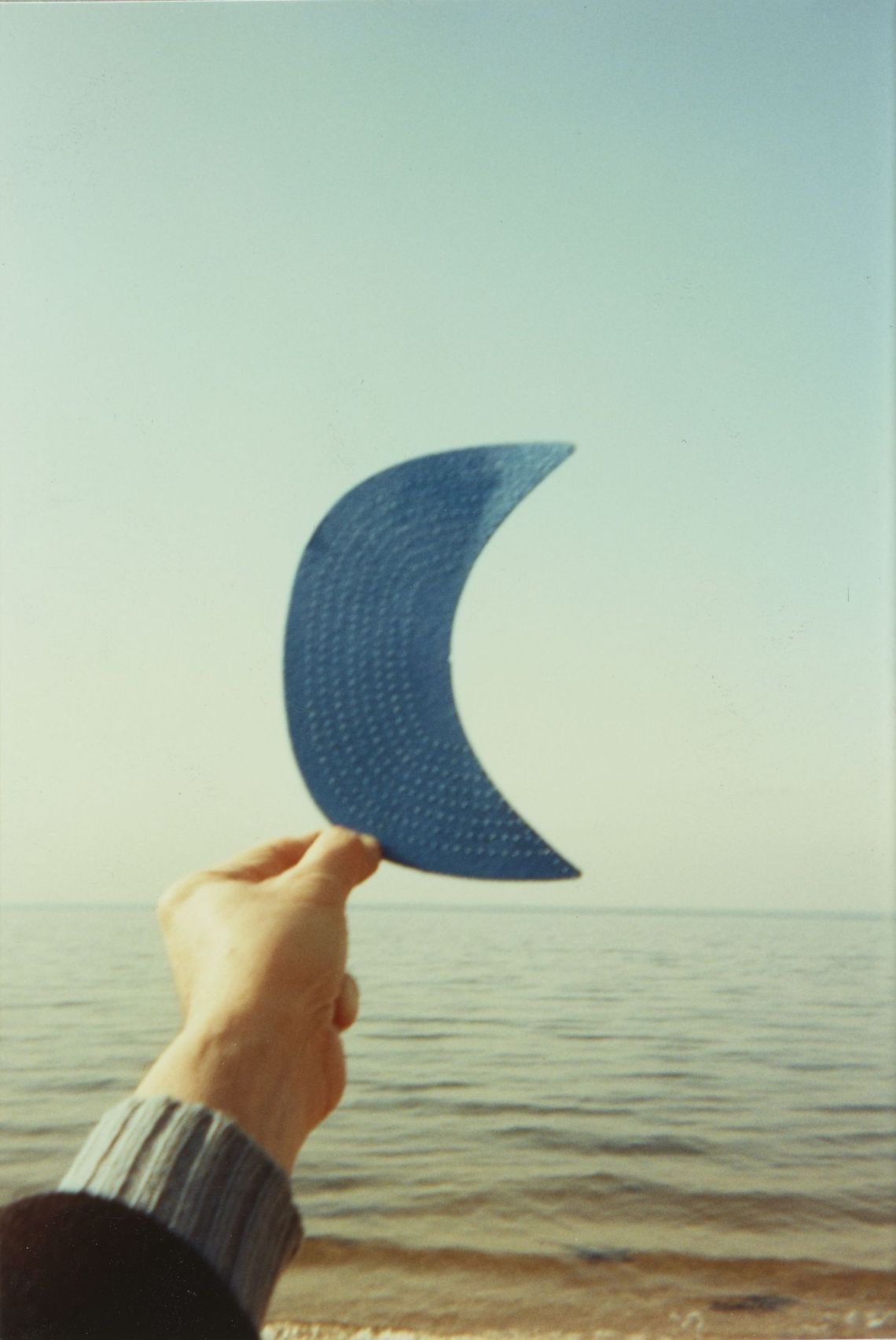 A hand holds a cutout of a crescent moon up to an ocean