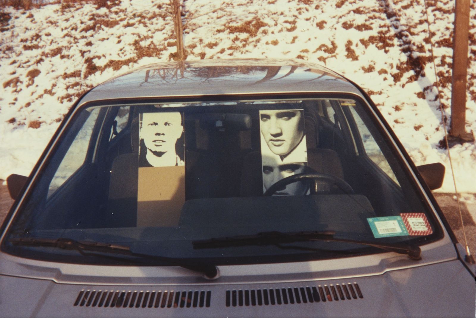 A parked car inhabited by three cut-out photographs, one of the artist and two of Elvis