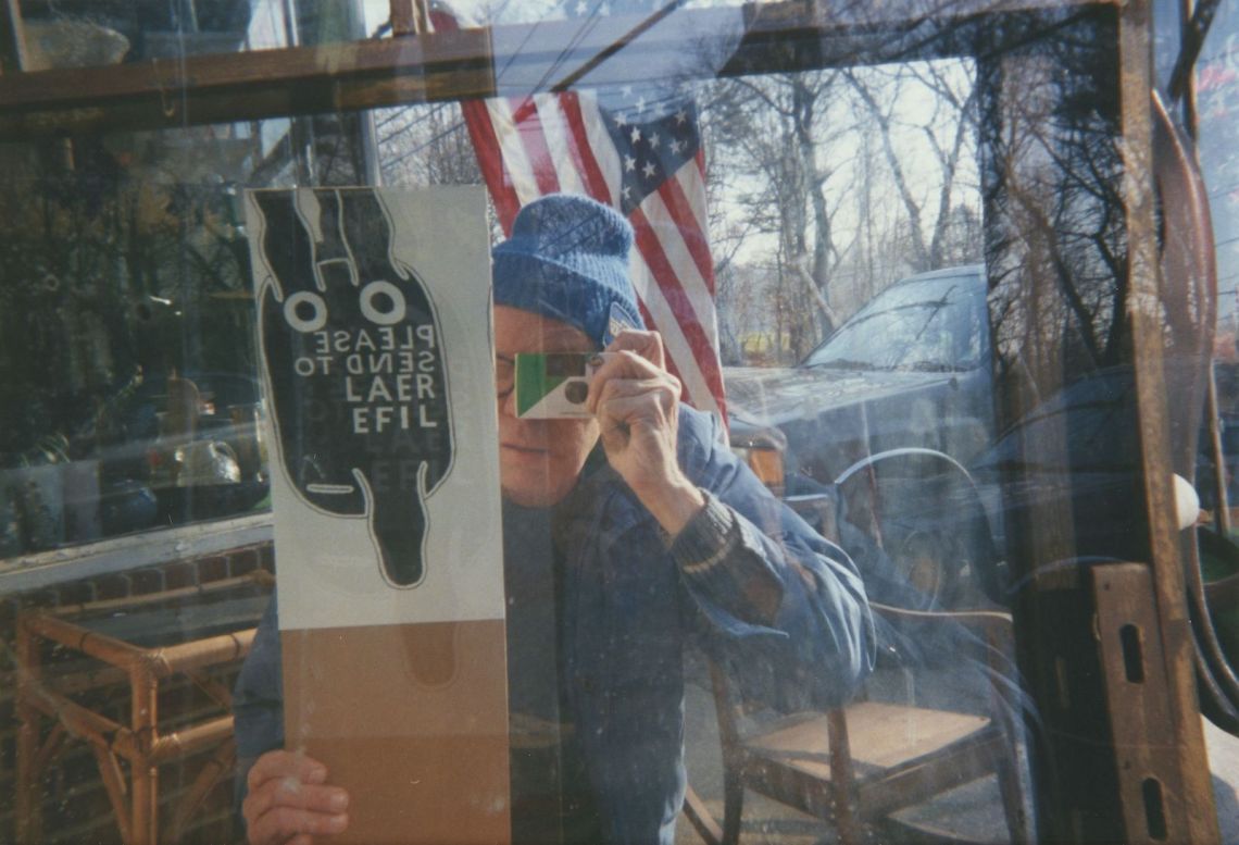 A middle-aged man taking a picture of his reflection in a window, holding a sign