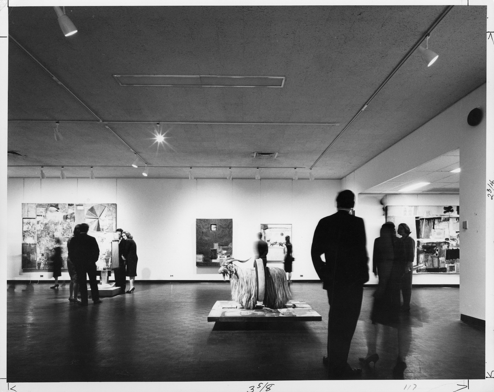 A black-and-white photograph of people swirling in a room with artworks on display