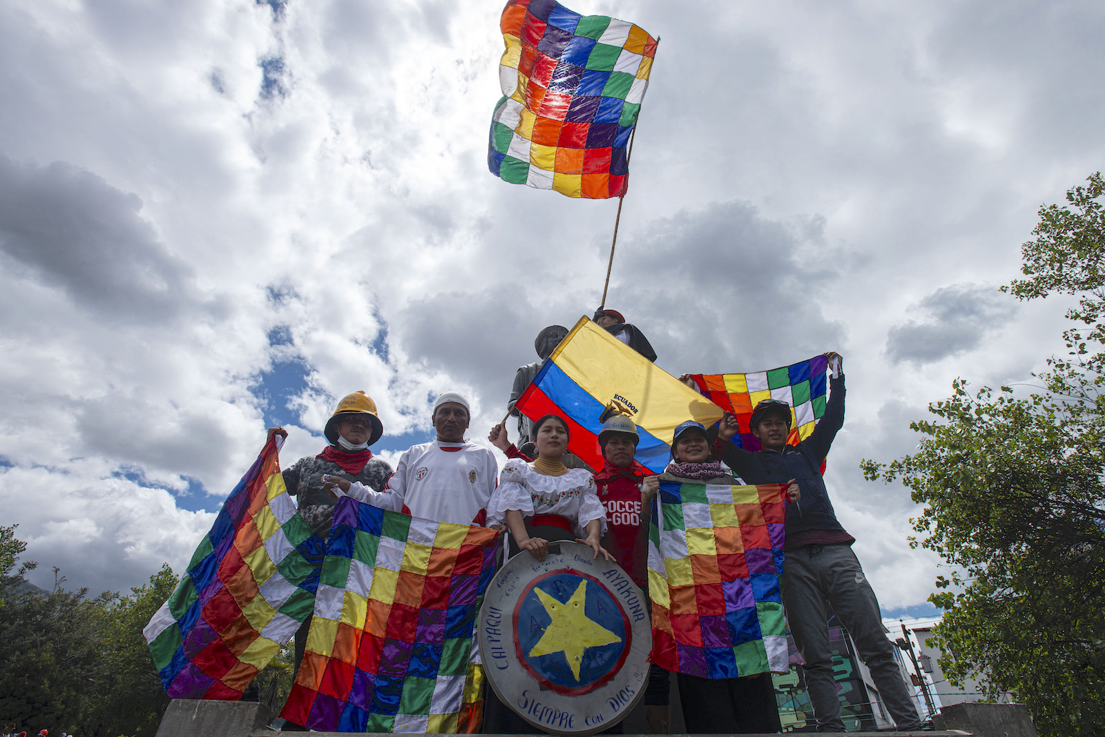 A group of protestors with rainbow checkered flags stand against a cloudy sky