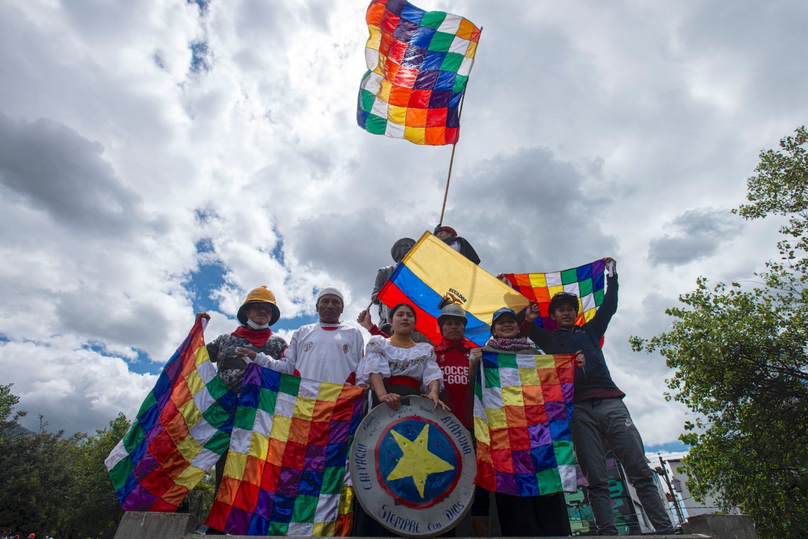 A group of protestors with rainbow checkered flags stand against a cloudy sky
