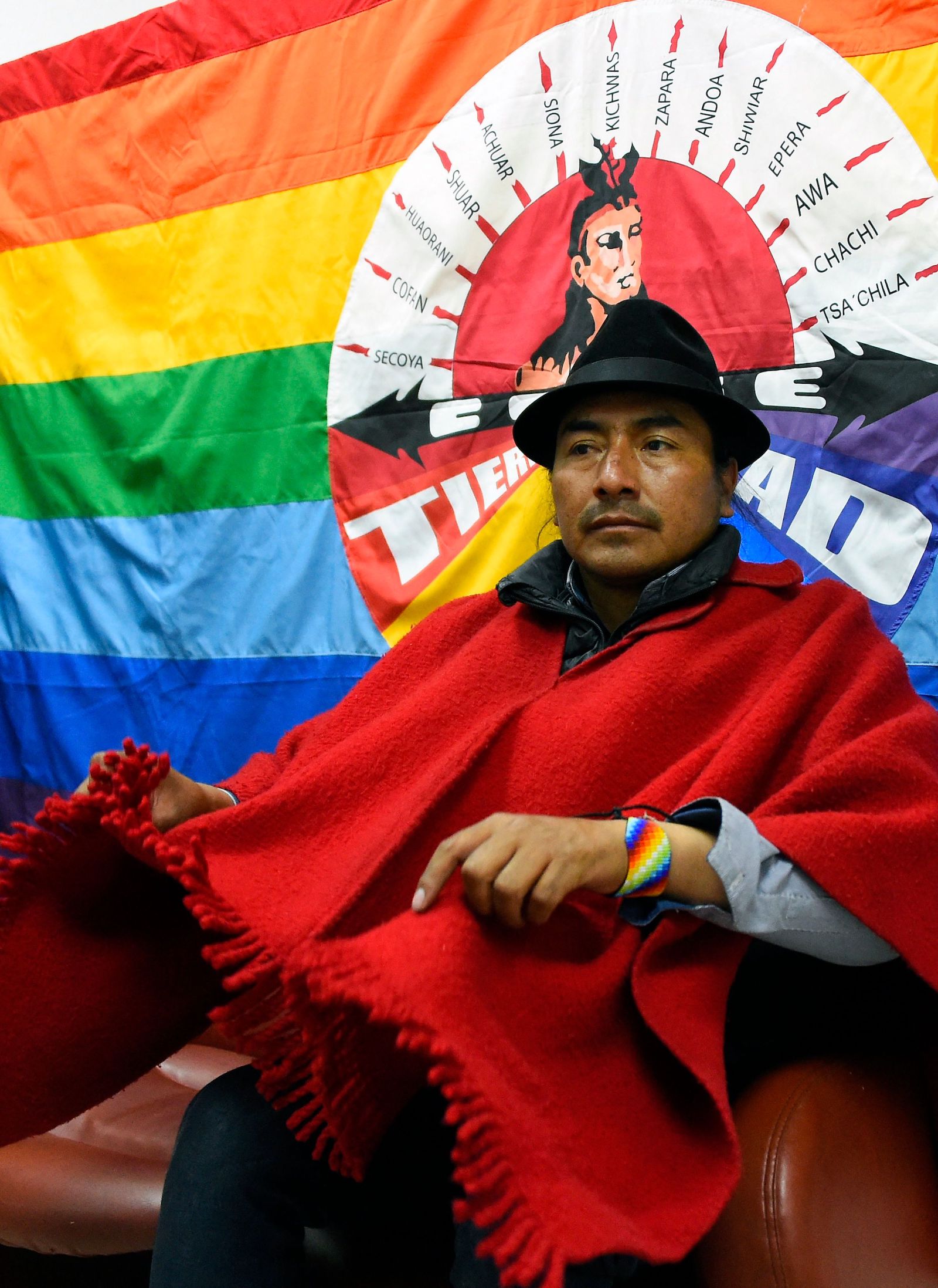 A man wearing red and a black hat sits in front of a rainbow flag