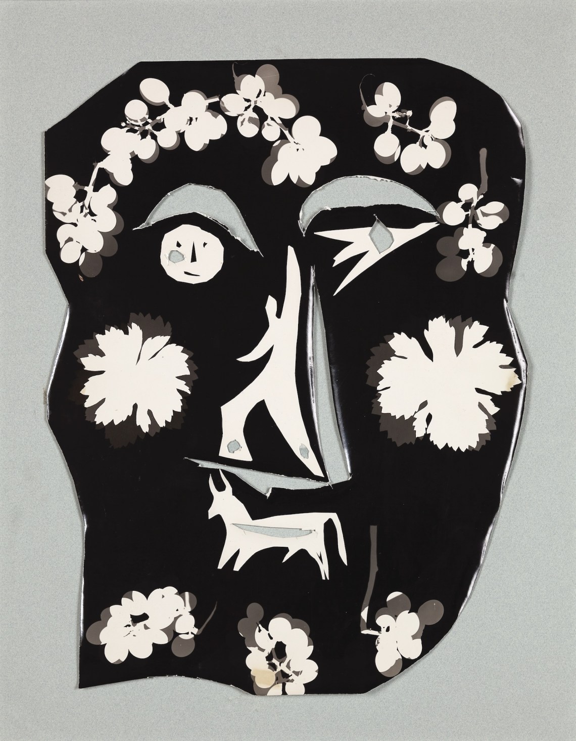 A black mask bedecked with white outlines of flowers, grapes, and animals, with a smiley face for the right eye