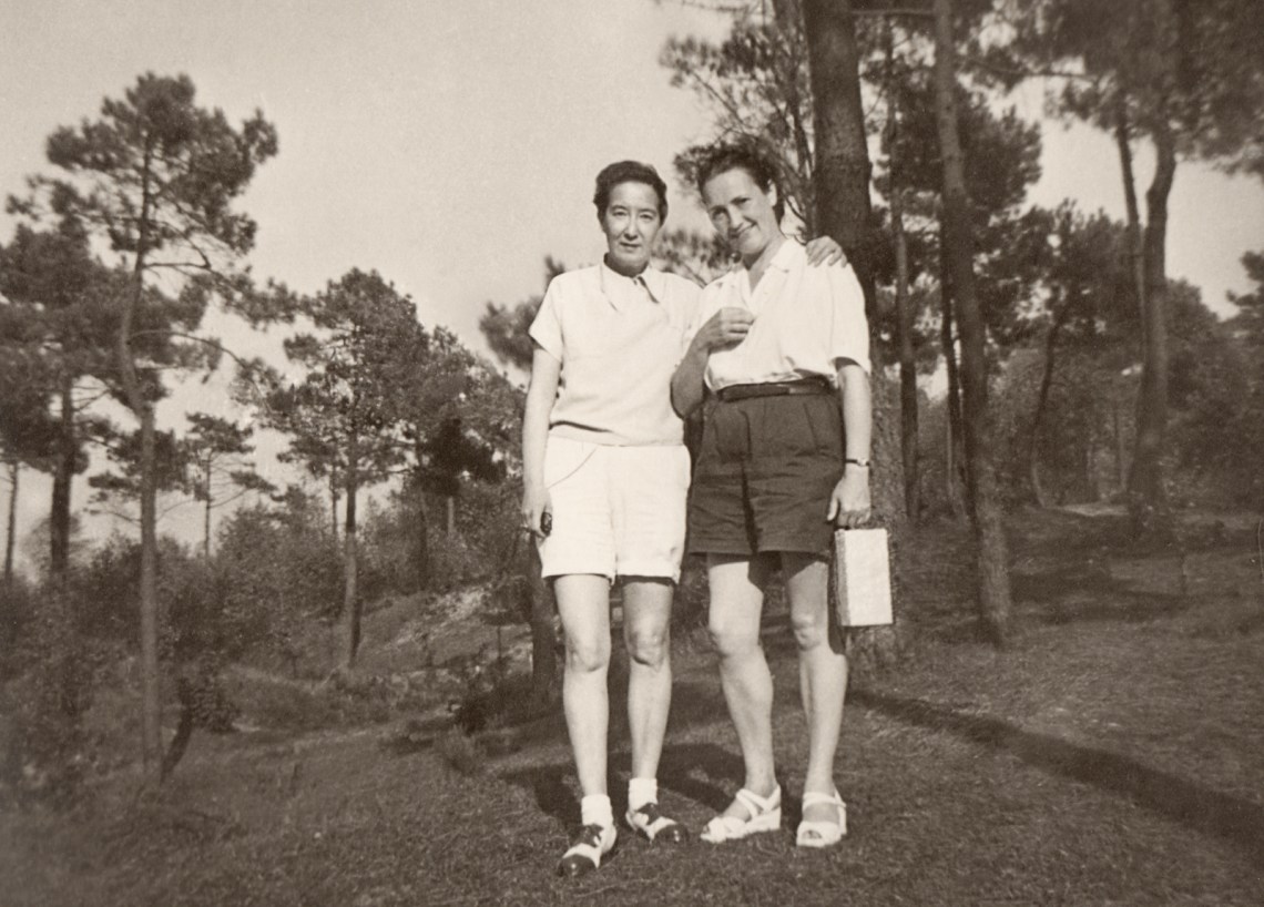 Two women posing in front of a forest, one with her arm around the other's shoulder