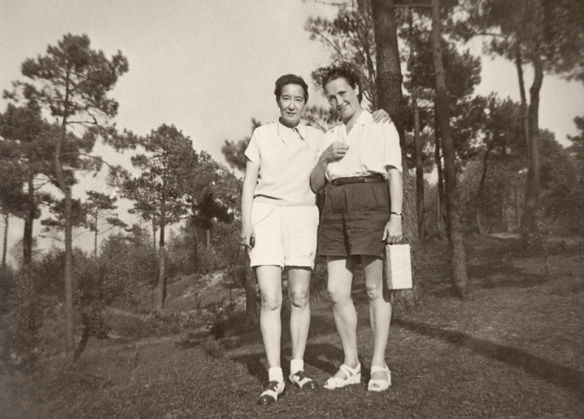 Two women posing in front of a forest, one with her arm around the other's shoulder