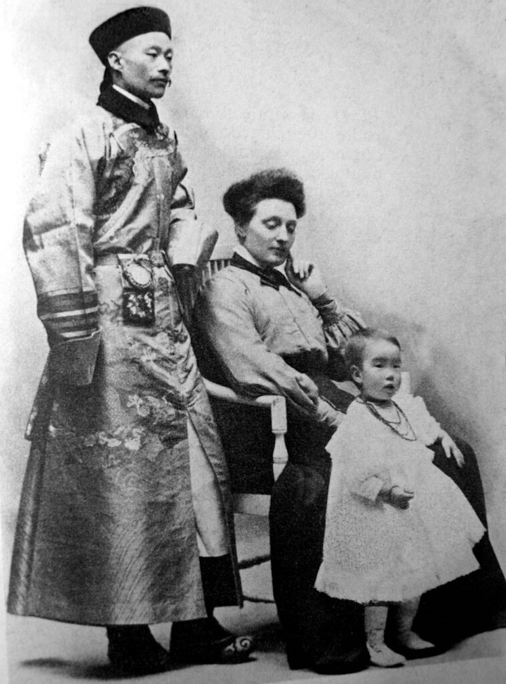 A portrait of a family in 1910s China, the husband standing on the left, his wife sitting midframe, his daughter standing on the right