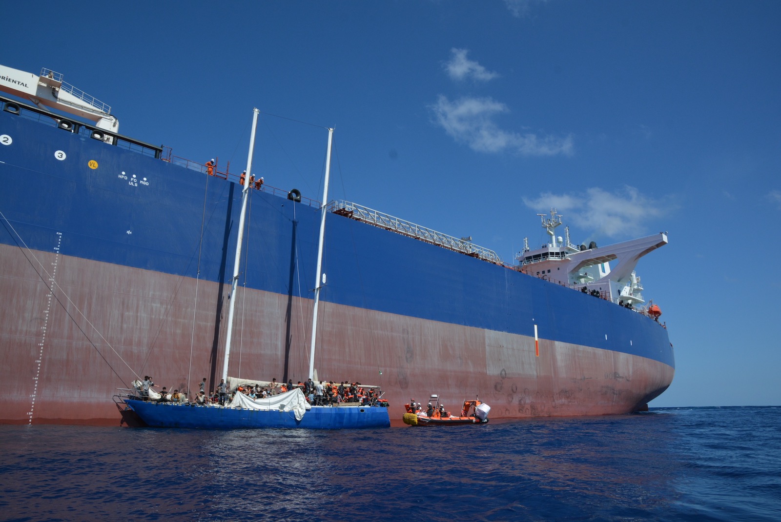 A small boat clinging to the side of an enormous grey and blue oil tanker at sea