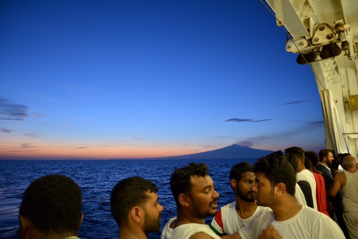 People gathered on the deck of a ship at twilight, the sea, the sky, and a mountain in the distance