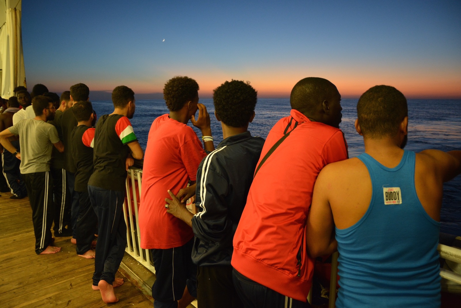 A row of people gathered on the deck of a ship, their backs to the camera, looking out at the sea at dusk