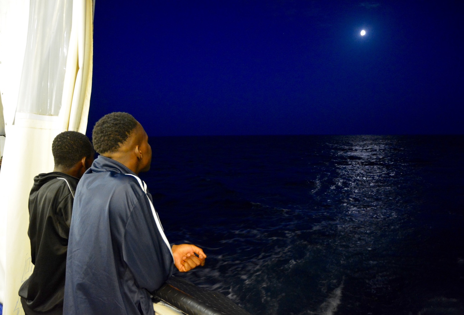 Two men look at the moon from the deck of a ship at night