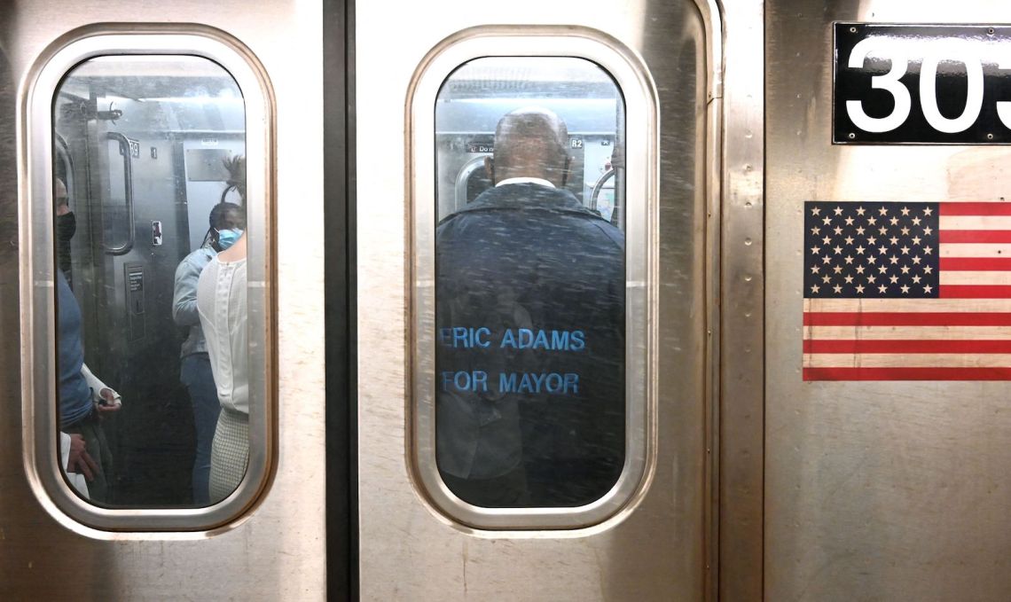 An exterior photograph of a subway car with an American flag on its side; inside the car stands a man with his back to the camera, wearing an 