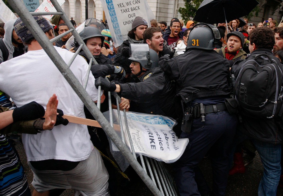 A struggle between police officers gripping a riot fence, and protestors, one of whom carries a sign, stuck in the fence, that reads 
