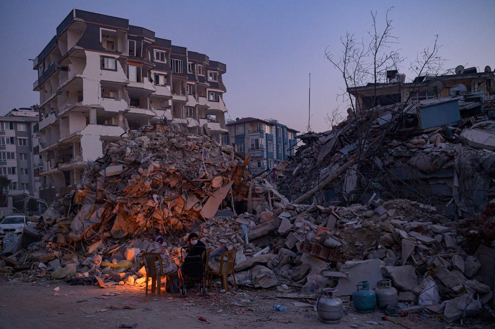 Two people sitting at a bonfire at twilight amid a pile of rubble, one building still standing behind them