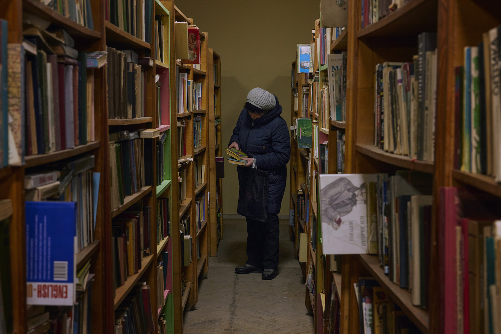 A woman in a gray hat and blue down coat holds books in an aisle between two walls of wooden bookshelves