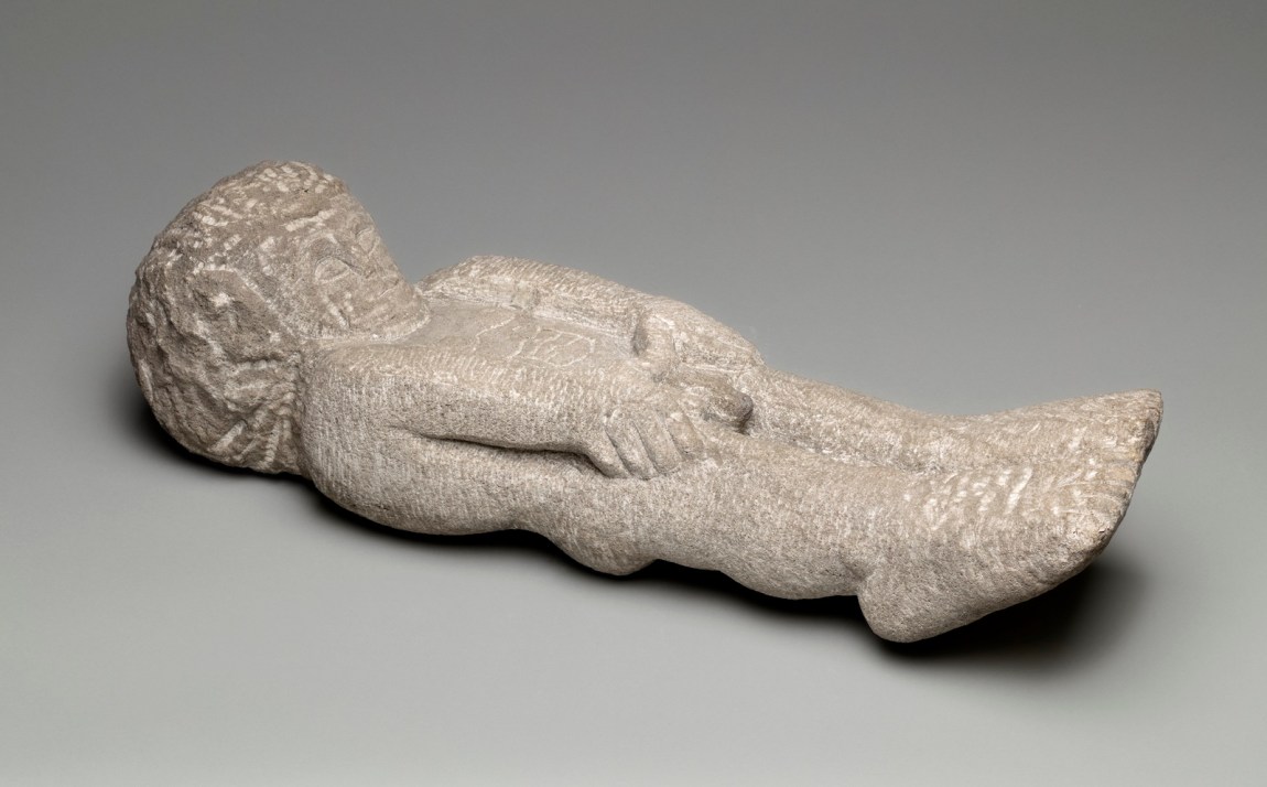 A carved limestone man lying on the ground with his hands on his thighs