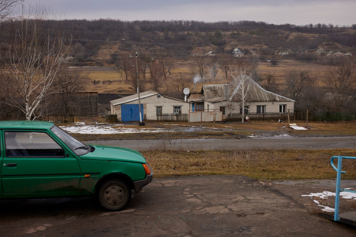 A cluster of buildings in a brown landscape on an overcast winter day, with some snow on the ground in patches, with a green car parked in the foreground