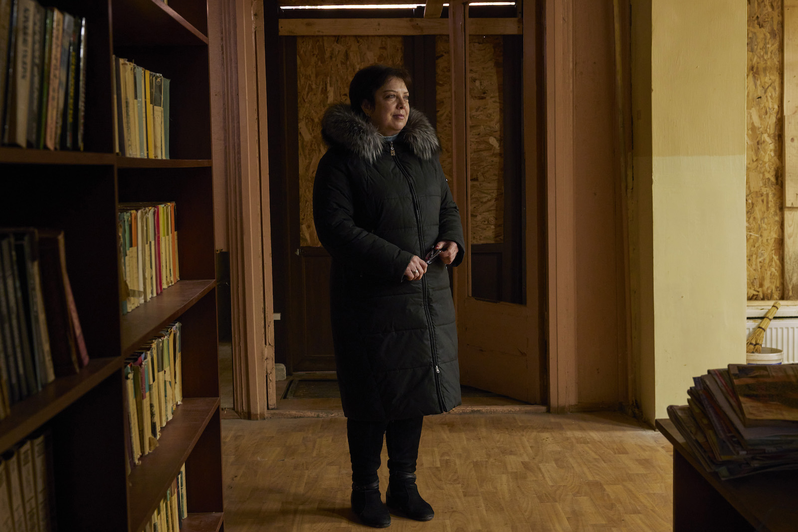 A middle-aged woman with short dark hair in a black coat with fur hood stands in a room with bookshelves and a door boarded up with plywood