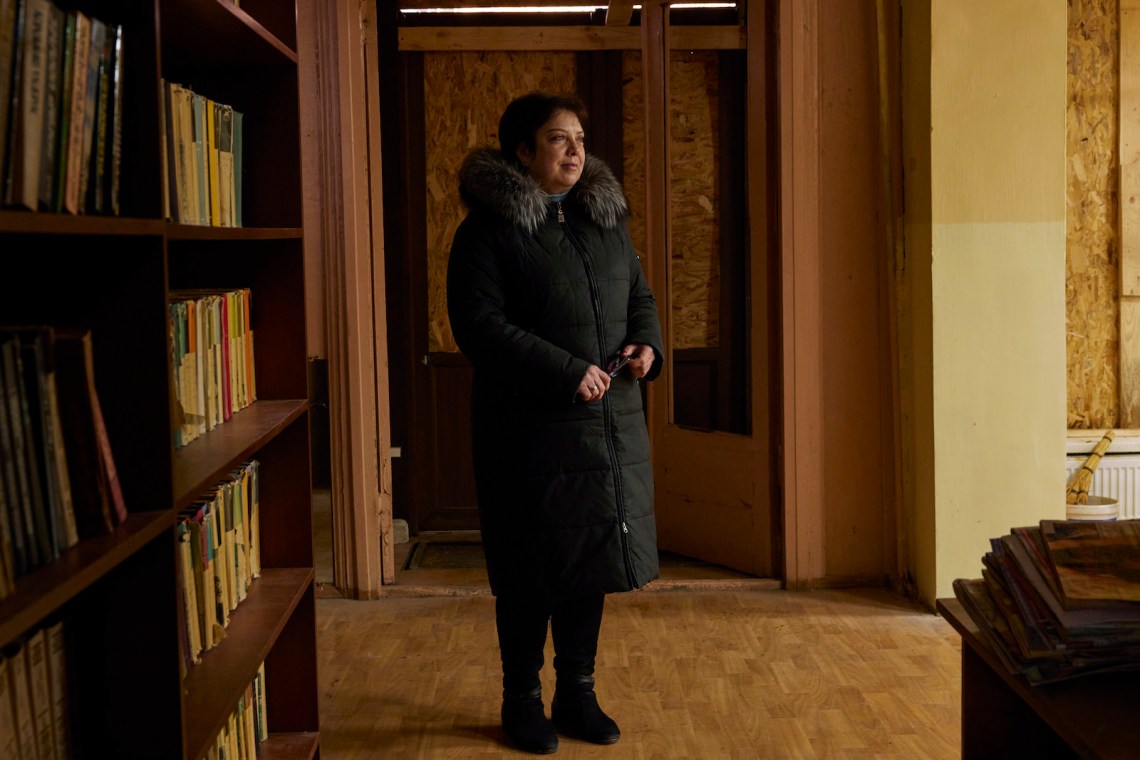 A middle-aged woman with short dark hair in a black coat with fur hood stands in a room with bookshelves and a door boarded up with plywood
