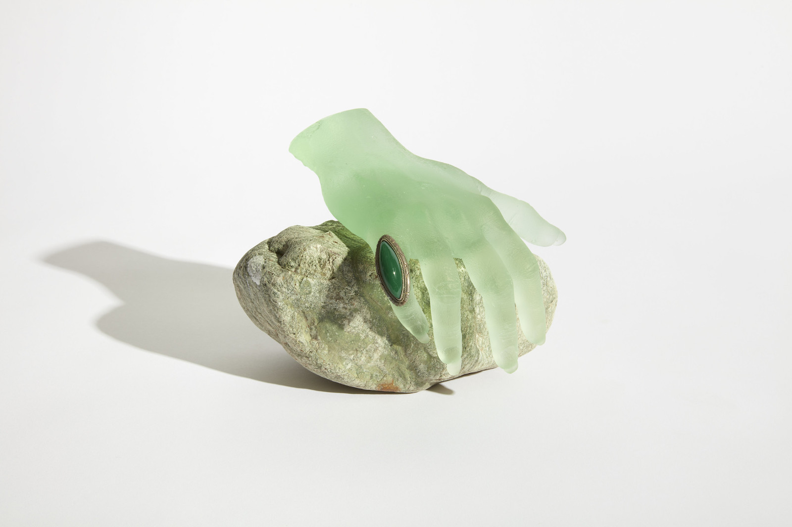 A frosted green glass hand wearing an oblong green ring rests on a stone