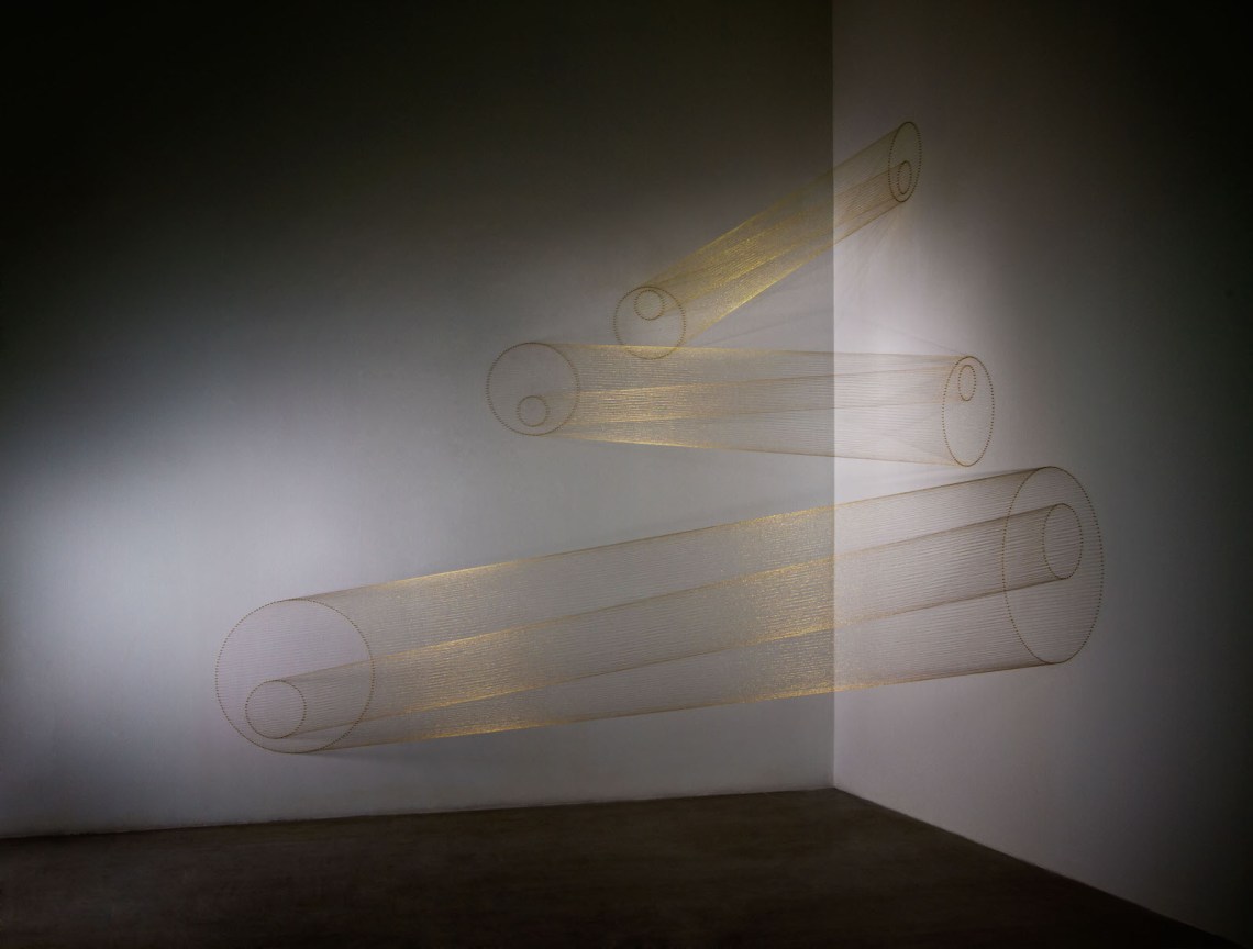 A sculptural installation in the corner of a gallery. Golden threads are strung between perpendicular walls to create the illusion of three floating, nested cylinders