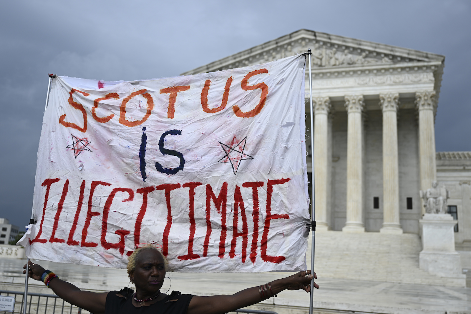 A woman holds a cloth banner in front of the Supreme Court that says SCOTUS IS ILLEGITIMATE in paint