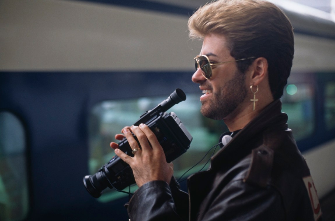 George Michael on a train platform holding a camcorder